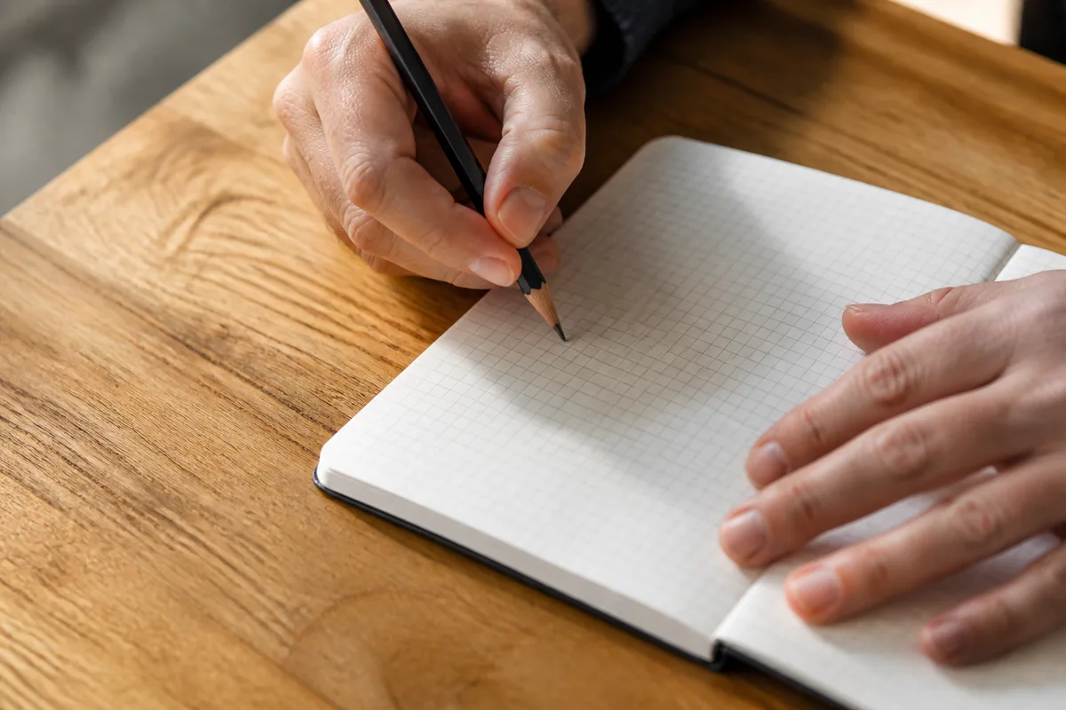 Mains annotant au crayon un carnet quadrillé posé sur un plan de travail en chêne